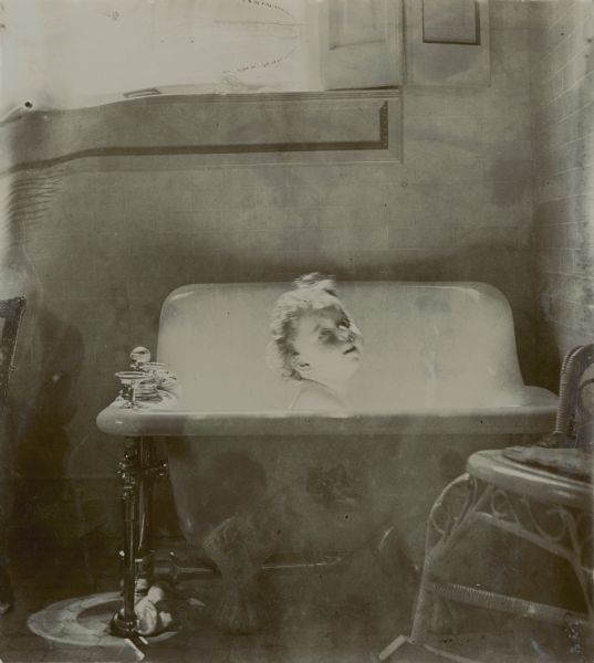 Sunlight through a window highlights the head and shoulders of Stuart Fargo, age 18 months, in a sitz bathtub in the bathroom of the family home. There is a wicker rocking chair on the right.  