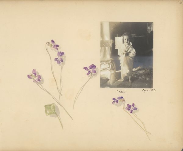 A decorated album page features a photograph of Stuart Fargo at age two years. He is wearing a nightgown and is barefoot. There is a wicker chair on the left and a table or dresser in the background. The photograph is captioned "nite" and also has the notation "2ys -1899." There are hand-painted violets and a violet leaf on the page.