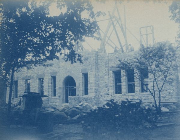 A cyanotype print of the Lorenzo Dow Fargo Library under construction.  Workmen are positioning roof trusses for the Gothic revival building. The stonework for the first story facade has been completed. There are thick stone lintels over the windows and the front door features a pointed arch. There are boulders and stone rubble in front of the building.