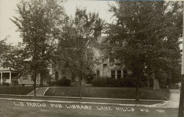 The Lorenzo Dow Fargo Public Library building is partially obscured by trees in this photographic postcard. The stone, Gothic revival building has a central spire. There are broad steps leading from the sidewalk toward the front door. The street is not paved. A two-story house with front porch is on the left. Caption reads: "L.D. Fargo Public Library, Lake Mills, Wis."