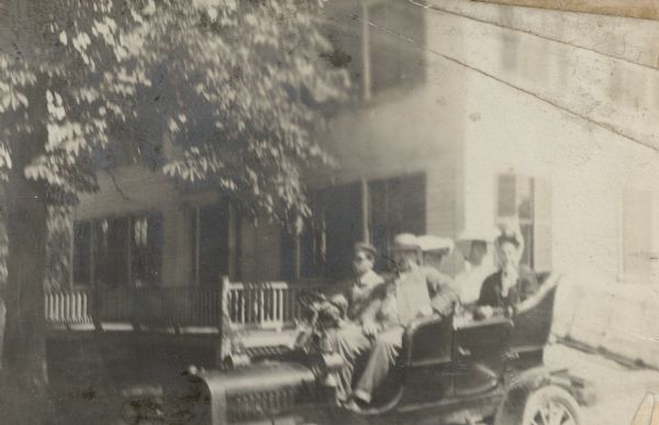 Five people posing in an automobile parked in front of a large frame house. The house is identified as the birthplace of Elizabeth Farnsworth Mears (1830-1907), an author and poet who lived most of her life in Oshkosh and published under the pen name Nellie Wildwood. Mears, wearing a dark dress, is sitting in the back seat with two of her daughters, sculptor Helen Farnsworth Mears and author Mary Mears. Frank B. Fargo, the husband of Louise Mears Fargo, is in the front seat, right. The driver is identified as Dr. [?Francis] Stanley, of Boston. A handwritten caption accompanying the photograph explains, "Grandma Mears left in emigrant wagon 1836, returned in automobile 1906."