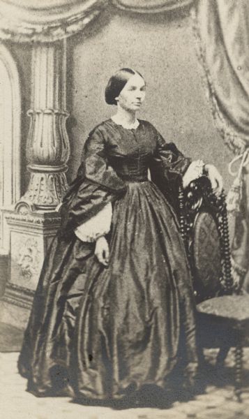 A photographic copy of an early, full-length studio portrait of Elizabeth Farnsworth Mears (1830-1907) standing in front of a painted backdrop with column behind her. She is wearing a dark floor length dress with full skirt and sleeves. There is light-colored trim at the neck and wrists. Her left forearm is resting on the back of a chair. Mears moved to Wisconsin as a child from her home state of Massachusetts and lived most of her adult life in Oshkosh. She was an author who published under the pen name Nellie Wildwood. Her three daughters were sculptor Helen Farnsworth Mears, author Mary Mears, and illustrator (<i>The Beautiful Land of Nod</i>) Louise Mears Fargo.