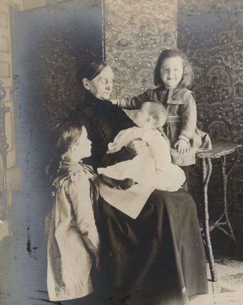 Elizabeth Farnsworth Mears, seated in front of a folding screen, holding her grandson Frank Barber Fargo, age six months, on her lap. Stuart Fargo, age three years, is smiling at the camera. He is sitting on a rattan table and has his right hand on his grandmother's shoulder. Older sister Dorothy, age six, is standing on the left.