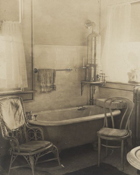 A wicker rocker and bentwood side chair flank the bathtub in a bathroom of the Frank Brown Fargo house. There is a hot water heater above the tub in the corner of the room and a steam radiator is on the right. The floor and walls are tiled. Two windows fill the room with light. A caption describes this as the "Bathroom off Mother's room."