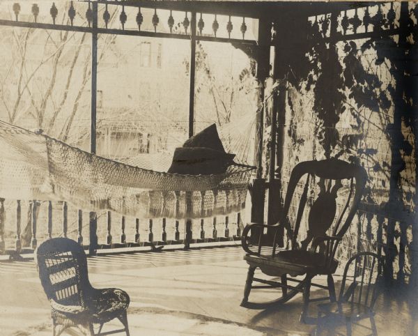 A photograph bearing the caption "Sleeping Porch off Mother's [Louise (Mrs. Frank) Fargo's] Room" depicts a screened porch with turned balusters and a spandrel with spindles. A rocking chair and two children's chairs are in the foreground. A hammock with pillows and fringed netting is hanging behind the chairs. There is foliage on the right. The large house next door, which belonged to Frank Fargo's brother, Enoch J. Fargo, is in the background. 