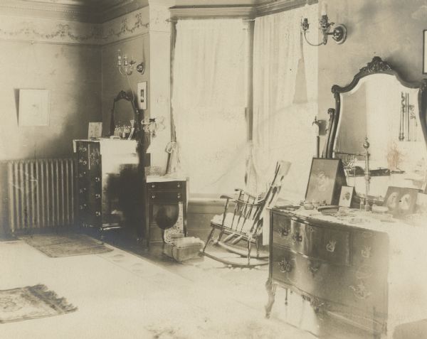 A highboy dresser, left, and its lowboy counterpart flank the large bay window in Louise Mears Fargo's bedroom. There is an electic wall sconce on either side of the window. The cord of a small lamp over the sewing cabinet to the left of the bay window is plugged into one socket of the sconce above it. There is a rocking chair and footstool in the bay. A carpet and several throw rugs cover the floor. There is a wallpaper frieze covering the area between the crown molding and picture rail molding. Portions of a brass bed are visible in the mirror over the lowboy dresser.