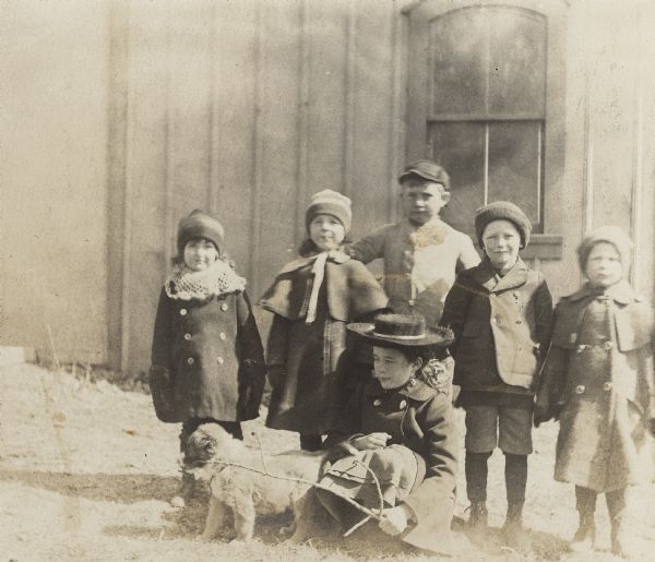Dorothy Fargo, center, is squatting down next to her puppy, Mark II.  Dorothy is wearing a winter coat and wide brimmed hat and is holding a stick. Five children are standing and posing behind her. They are, left to right, her two brothers, Frank and Stuart; William Griswold; Philip and Dell Falk. They are standing in front of a carriage house.