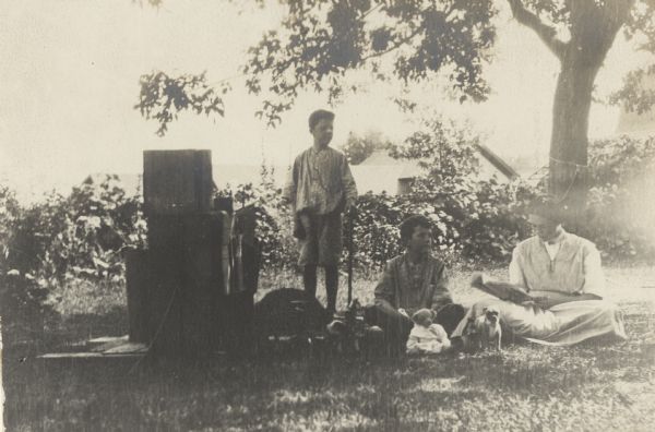 The three Fargo children, from left, Stuart, Frank Jr. and Dorothy are posing with a toy barn made of wooden boxes. Dorothy is reading a magazine. There is a teddy bear and small dog on the ground in front of Frank.  