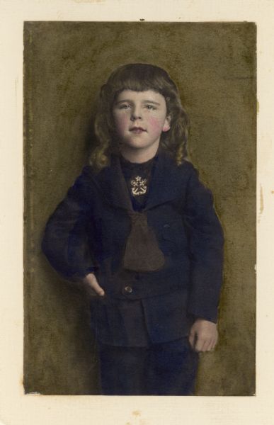 A standing, hand-colored three-quarter length portrait of Stuart Fargo. He is wearing a jacket over a shirt with an embroidered design at the neck. A caption written below the photograph reads: "6 yrs - just before hair was cut."