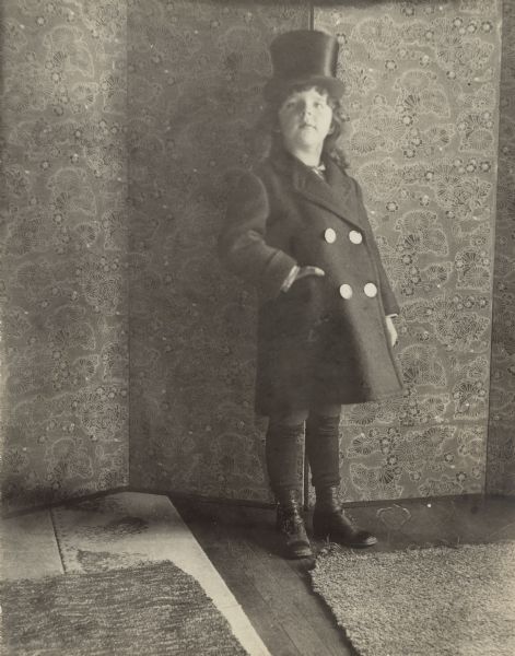 A six-year-old Stuart Fargo is posing for a full length portrait dressed as the "New Year" while standing in front of a folding screen. He is wearing a top hat, heavy double breasted coat, and high-buttoned shoes. There are three rugs on the floor.