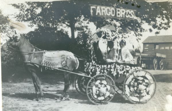 Two unidentified boys pose in a horse-drawn carriage with a banner above it which reads: "Fargo Bros." The open carriage has an umbrella top and is decorated with paper streamers. The horse harnessed to the carriage is wearing a fringed fly-net and has decorations over its ears and tail. There is a shed and wagon in the background.