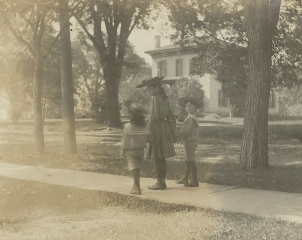 Dorothy Fargo, ten years old, is holding the hands of her brothers Frank, four years old, and Stuart, seven years old, as they are posing on the sidewalk along Mulberry Street near their home. Stuart is looking back at the photographer. A brick Italianate house is on the opposite side of the street. A handwritten caption notes that the photograph was taken on the "Boys first day at Kindergarten."