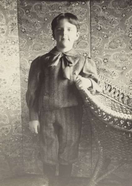 Stuart Fargo posing for a three-quarter length portrait standing in front of a folding screen with his left arm resting on the back of a wicker chair. He is wearing a heavy shirt with matching trousers that reach below the knee, and a large bow tie. In photographs up to this age Stuart has had long hair.