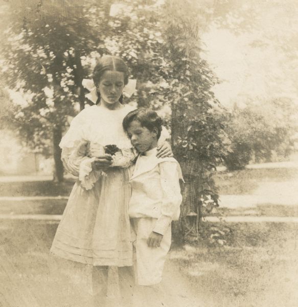 Dorothy Fargo is holding a flower in her right hand and is resting her left hand on her brother's shoulder as they are posing outdoors for a three-quarter length portrait. She is wearing a light-colored dress and a large bow in her hair. Stuart is wearing a sailor suit. There is a trellis or arbor behind them.