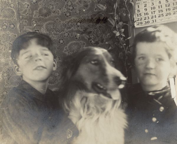 Stuart, left, and Frank Fargo posing with their dog, Mark, in front of a folding screen. There is a calendar on the screen behind them.  