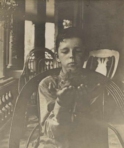 Thirteen-year-old Stuart Fargo posing on the front porch of his home holding a wild bunny. He is sitting on a bent willow chair; two other chairs are in the background.