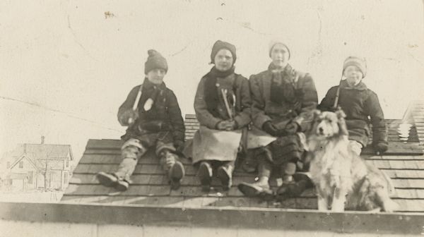 Four children, dressed for cold weather, posing while sitting on the ridge of a roof. They are, from left: Frank Fargo, Florence Anderson, Dorothy Fargo and Stuart Fargo. The Fargo family dog, Mark, is sitting in front of the children. There is a house in the background on the left.