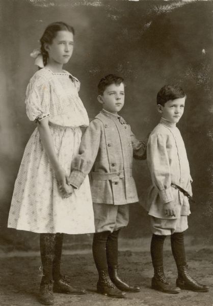 The three children of Frank B. and Louise Mears Fargo standing and posing for a full-length studio portrait. They are, from left: Dorothy, age 12 years; Stuart, nine, and Frank six. The boys are wearing suits with short pants and high buttoned shoes. Dorothy is wearing a light-colored print dress and a bow in her hair.