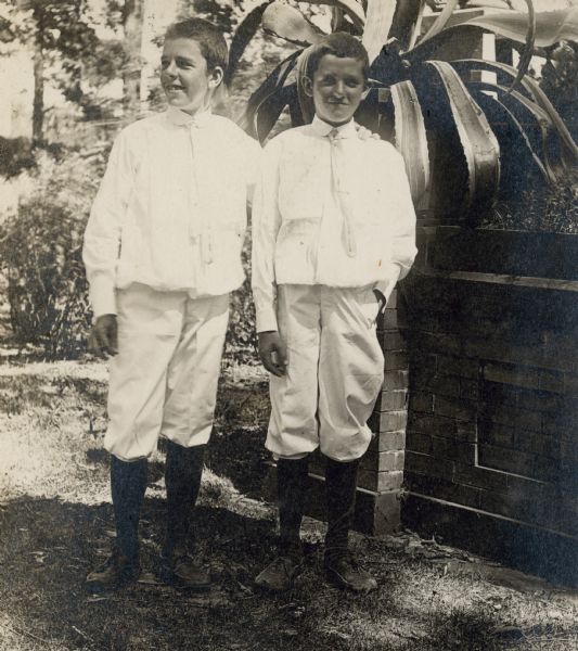 Brothers Stuart, left, 15 years old, and Frank Fargo, 12, posing in front of the front porch of the family home. There is a large potted agave on the porch wall. The boys are wearing light-colored shirts, short pants and neckties.