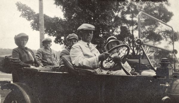 Frank B. Fargo, wearing a double-breasted duster and cap, is sitting at the wheel of an open automobile. His wife, Louise Mears Fargo, is sitting beside him. In the back seat, from left, are: Mrs. (?Charles) Beveridge, Frank Fargo Jr. and Stuart Fargo. A handwritten caption identifies the location as Lake Geneva.