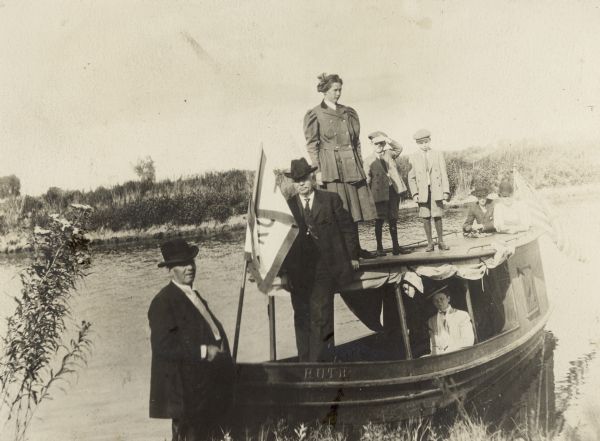 Frank B. Fargo, standing on the shore and posing in front of the small launch "Ruth" on Rock Lake. An unidentified man, possibly Charles Millard, is standing on the lower deck; Fargo's three children, Dorothy, Frank Jr. and Stuart are standing on the upper deck. There is a woman in the cabin and two men standing at the rear of the boat leaning on the upper deck.  