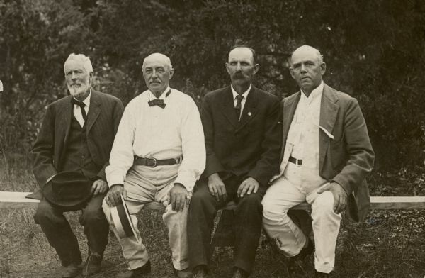 Frank B. Fargo of Lake Mills, far right, posing, sitting on a board, with three other well-dressed men at the Holstein Friesian Association Picnic. The other men, as identified by a handwritten caption, are, from left: Wm. Everson, F.S. Greenwood, and M. DeMerett.  