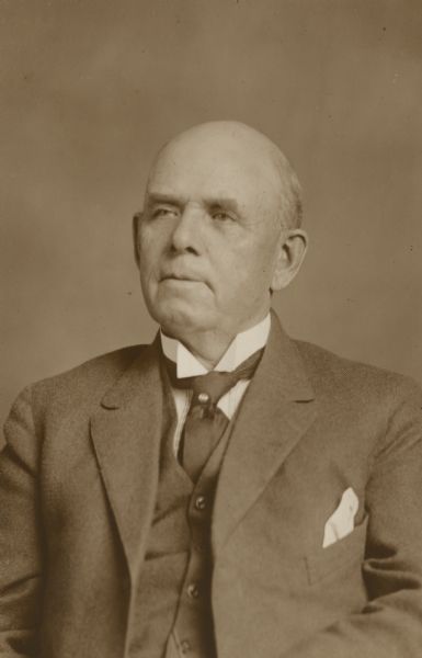 A seated head and shoulders studio portrait of Lake Mills businessman Frank B. Fargo at age 68. He is wearing a suit with vest and necktie with stickpin. There is a handkerchief in his jacket pocket.