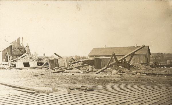 A large section of roof is on the ground in the foreground, sections of timber framing have collapsed, and exposed framing on a standing section of a building is on the left. An intact barn with two roof ventilators stands in the far background. The damage is from a "cyclone" on the Frank B. Fargo farm.