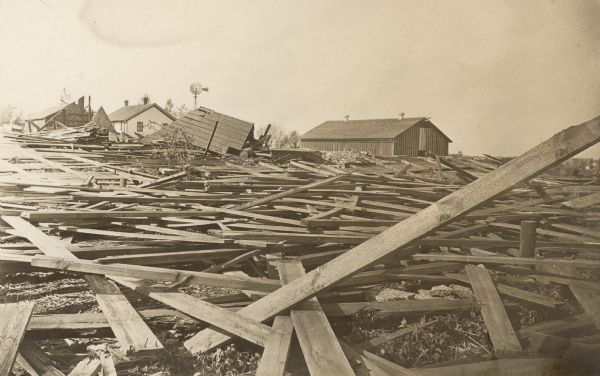 Boards are scattered on the ground after a "cyclone" on the Frank B. Fargo farm. There is a large section of roof on the ground. In the background is a section of a barn with exposed framing, a portion of the apparently undamaged farmhouse, a windmill, and a large barn with two roof ventilators.