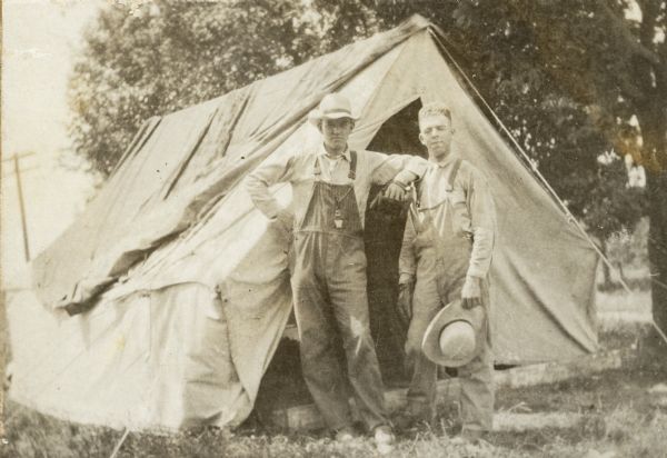 Frank Fargo Jr. is resting his elbow on his older brother Stuart's shoulder as they are posing standing in front of a large tent. Both are wearing bib overalls; Frank is wearing a hat while Stuart is holding his. Frank sports a watch fob hanging from his overall breast pocket. A handwritten caption notes that the photograph was taken on the family farm.