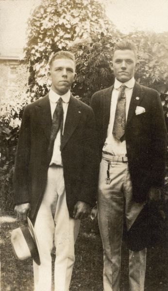 Stuart Fargo, left and his younger brother Frank Fargo Jr. pose for a three-quarter length portrait outdoors. Both men are wearing suits, and ties with lapel pins. Both men are holding their hats. Frank has a watch fob in the shape of the letter W hanging from his pants pocket.