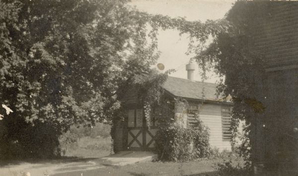 Vines grow on the walls of a small wood frame building with double doors on the gable end. A sloped concrete slab leads to the doors. A handwritten caption identifies this as the "Home Garage" of the Frank B. Fargo house on Mulberry Street. There are wooden boards partially covering the windows and a ventilator on the roof. The corner of another building is in the right foreground.