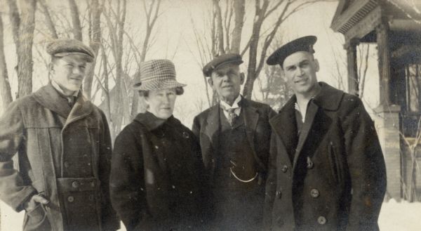 From left, Frank B. Fargo, Jr. posing with his parents, Louise Mears and Frank B. Fargo, and Edw. Webster near the porch of the Fargo family home. Webster is wearing a Navy uniform with hat and pea coat; the others are also wearing winter clothing. There is snow on the ground.