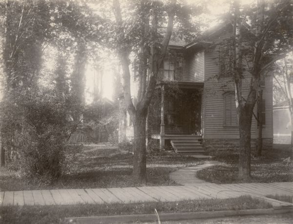 View from street of a two-story wood frame house, with open front porch shaded by trees. There is a plank sidewalk in front and a curved plank walk leading to the front steps. It is identified in a handwritten caption as "Mother's home Oshkosh Wis — Polk St. where she lived from 9 yrs. until she married 1893." "Mother" refers to Louise Mears Fargo, the daughter of (Mary) Elizabeth Farnsworth Mears and John Hall Mears. The Mears' had two other daughters, sculptor Helen Farnsworth Mears and author Mary Mears. Elizabeth Mears was a poet whose work was published under the pen name Nellie Wildwood. Polk Street has been renamed Parkway Avenue.  
