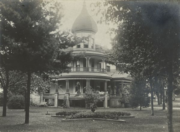 A corner view of the Frank B. Fargo house is centered on the onion-domed polygonal tower. The open, wrap-around two-story porch features paired columns, balustrades, and a spandrel on the second level. Two hammocks are hanging on the porch. There is a star-shaped flower bed in the foreground.  