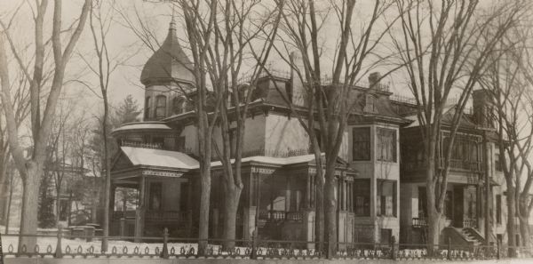 A view of the side and front of the Second Empire style, two and one-half story cream brick house on Mulberry Street. There is an onion domed tower on the left; the front porch extends around the corner of the house as a screened porch. There is also a second story screened sleeping porch above the side entrance and a two-story bay on the side. Three chimneys are visible in this view. In the foreground is a low stone or concrete wall topped with an open ironwork fence. In the background on the left is the home of Robert Fargo, the uncle of Frank B. Fargo.