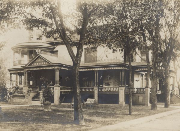 View from street of Fargo house on Mulberry Street. A hammock and chair are on the open front porch. Foliage obscures the top of the tower on the left, but its windows are visible above the porch roof. There is a bent wood settee on the lawn; a large urn on a pedestal holds potted plants. The porch has paired columns and ornate decorative trim.