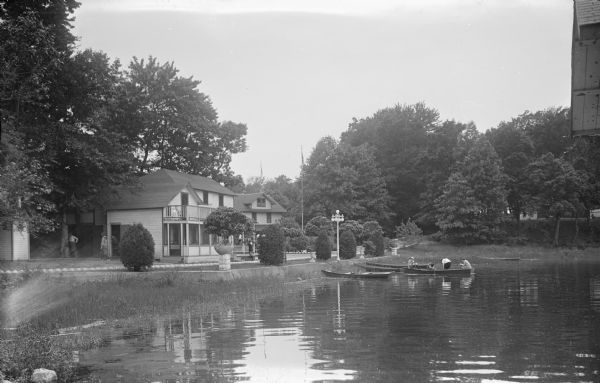 View across water towards a section of Sandy Beach Resort. There are people on the lawn on the left next to buildings. Two women are talking on a porch in the center, and four people are sitting in a boat near three boats on the shoreline.