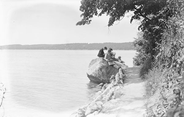 View along shoreline towards a group of people sitting on a large rock at the shoreline of the lake. Trees and plants are along a trail on the right. The far shoreline is in the background.