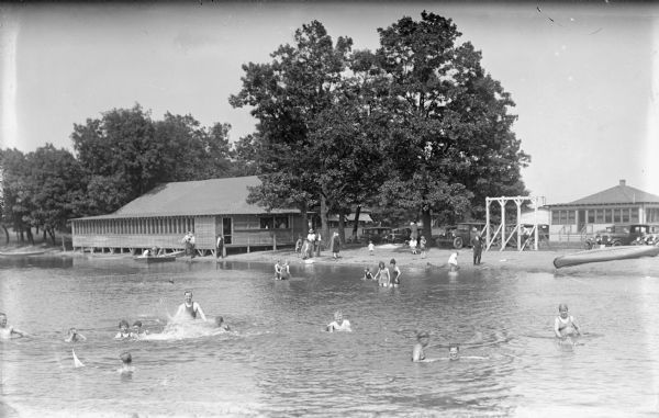 View across water towards shoreline. A large group of children and adults are swimming and playing in the water. There is a large building on the shoreline on the left, and more buildings on the right. Automobiles are parked nearby. People are standing, sitting and walking on the beach.
