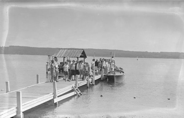 View from shoreline of a large group of people on a pier. Men and women are standing on the pier, posing and looking toward the shoreline. Other people are sitting underneath a roof at the end of the pier near a diving platform. Other people are sitting in a boat tied to the side of the pier. The far shoreline is in the background.