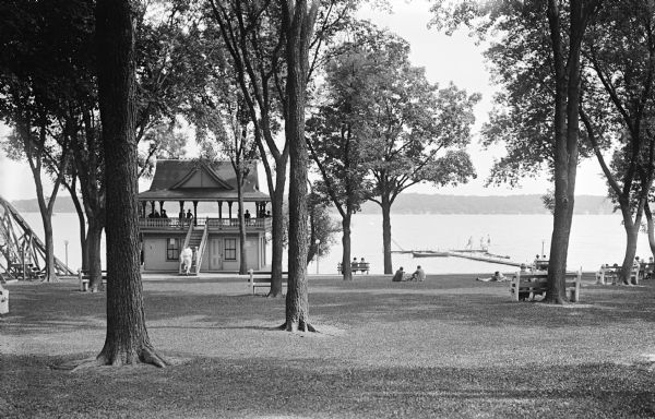 View across lawn towards a boathouse on a lake. On the far left is what appears to be a tall, wooden water slide, with a set of steep steps. People are sitting in a gazebo area above the boathouse, and sitting on benches on the lawn. There are boats moored to a pier on the right, and a group of people around a diving board. The far shoreline is in the distance.