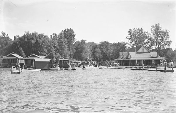 View across water towards shoreline filled with piers, boats, people and cottages. There is a large building on the right, and a parking area filled with automobiles.