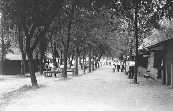 View of groups of people sitting at picnic tables under rows of trees that lead down to a lake at a resort. More people are standing or walking near buildings in rows on the left and right. On the right are small storefronts that have raised wood awnings with signs that read: "Check Room," and "Leave Your Films Here." There is a board on display with photographs near a tree. A lake is in the background beyond trees.