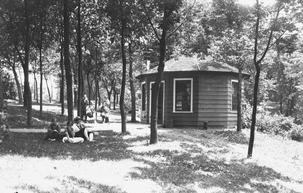 View of women and girls sitting on the lawn under trees near an octagonal building. They are all eating what appears to be ice cream.