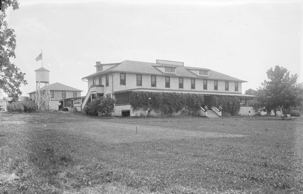 View across lawn towards a large building with a roofed porch. The front of the porch is covered heavily with vines. On the lawn in the foreground is a mowed area with stakes at either end, perhaps for croquet or horseshoes. On the left behind the building, a man is standing near a water tower that has a flag on a flagpole extending from its roof. Other buildings are in the background.