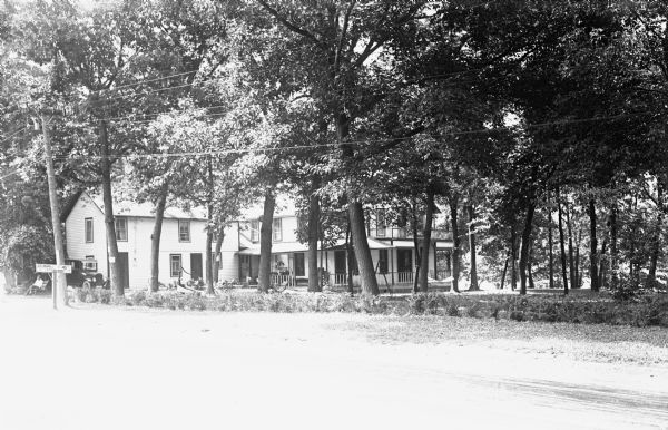 View from road towards a large, L-shaped building. There are benches and a hammock in the yard, and a hand-pump with a trough. A man is sitting on a bench on the far left near an automobile. A sign at the side of the road reads: "Ice Cream, Candy, Cold Drinks."