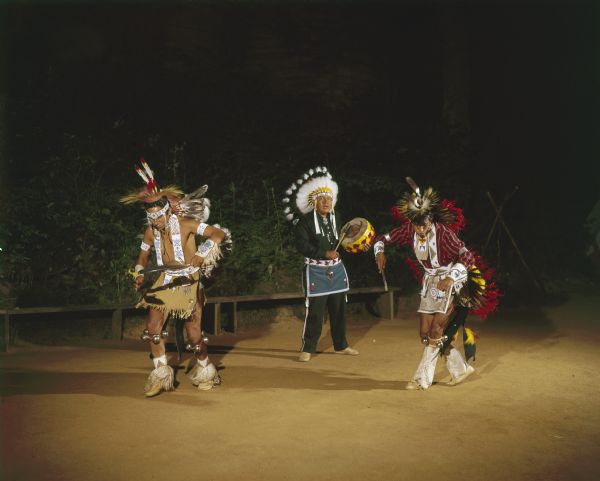 Two men and a drummer, with ceremonial clothing and headdresses, participating outdoors in the Stand Rock Indian Ceremonial Dance Challenge.