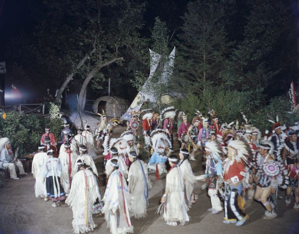 Elevated view of a large group of men and women in ceremonial clothing, dancing the Green Corn Dance of the Winnebagos. A sign in the background reads: "Sioux Tepee," and a tepee and what may be a chipoteke are set up under trees. People are walking and watching from a wooden walkway in the background on the left.