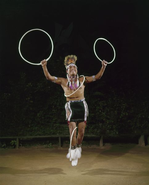 A male hoop dancer (Tony White Cloud) dressed in ceremonial clothing is jumping in the air, holding a hoop in each hand, and one hoop over one of his shoulders and across his torso, and a fourth hoop caught between his feet. 